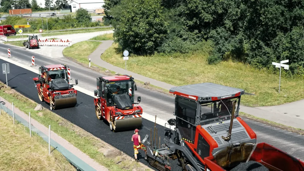 Three road rollers following a paver along a newly surfaced road approaching a junction